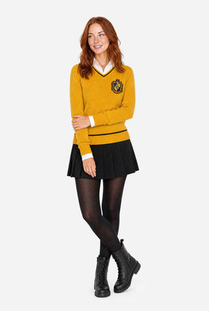 A woman modeling a mustard yellow long-sleeve Harry Potter Hufflepuff sweater with a black and white V-neck collar, a black crest patch on the left chest, a black pleated mini skirt, black tights, and black lace-up boots, set against a solid light gray background.