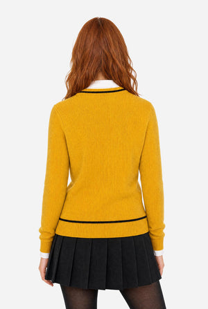 A woman with red hair is shown from the back wearing a mustard yellow ribbed knit sweater with black trim, a black pleated skirt, and black tights against a light gray background.