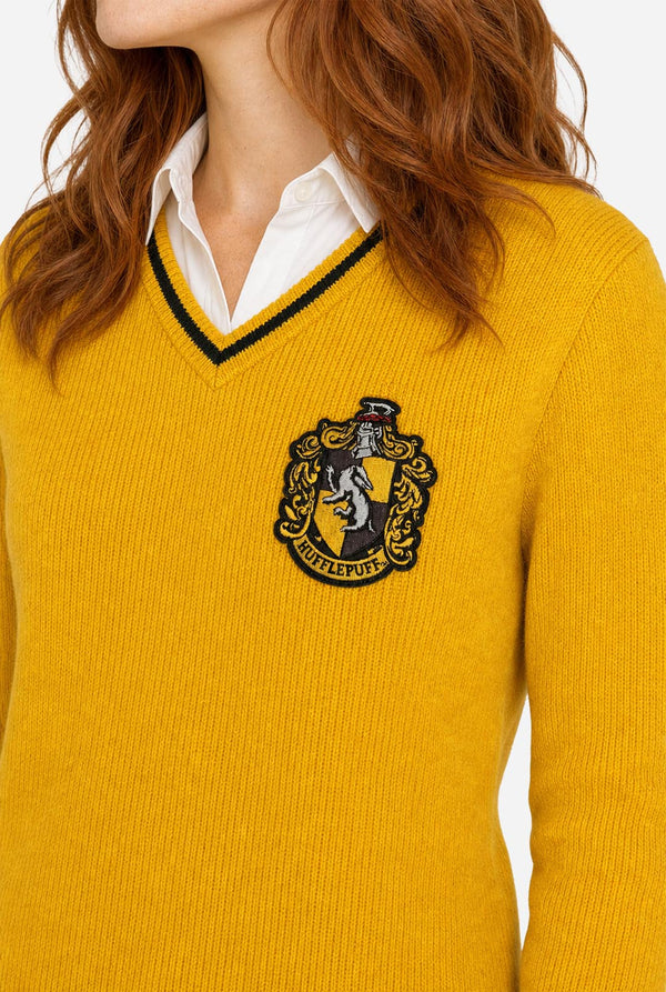 A close-up of a woman wearing a mustard yellow ribbed knit Harry Potter Hufflepuff sweater with a black and yellow V-neck trim and an embroidered Hufflepuff crest on the chest, layered over a white collared shirt.