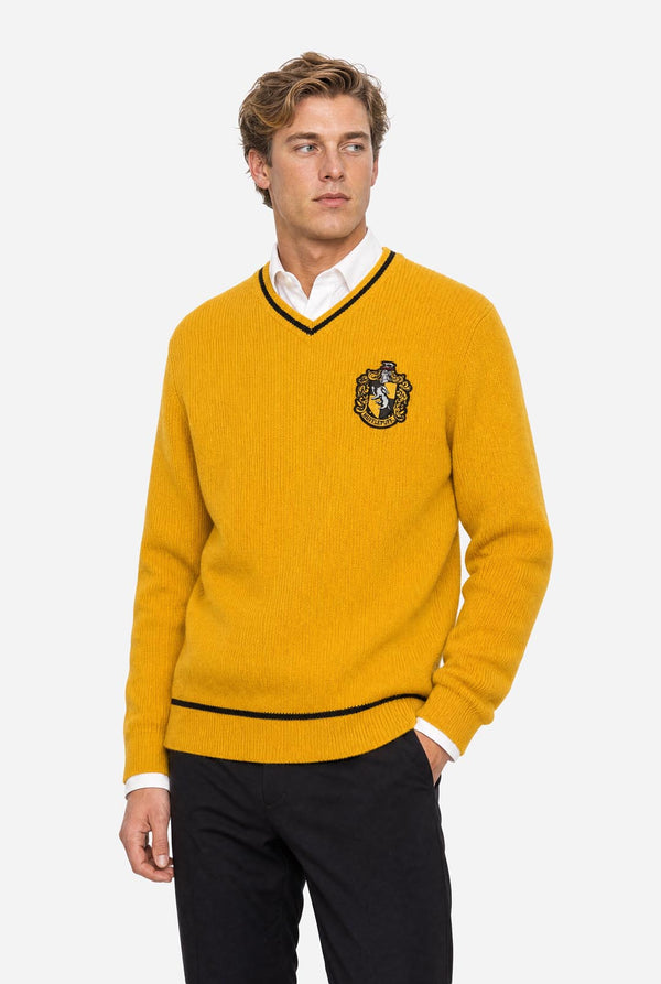 A man wearing a mustard yellow v-neck Harry Potter Hufflepuff sweater with black and white trim and a Hufflepuff crest on the chest, layered over a white collared shirt and paired with black trousers, against a light gray background.