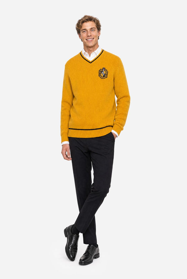 A man modeling a mustard yellow V-neck Harry Potter Hufflepuff sweater with black trim and a crest patch on the left chest, worn over a white collared shirt with black trousers and black shoes, against a light gray background.