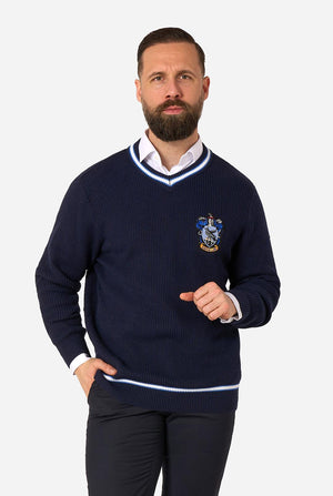 A man wearing a navy blue knitted V-neck Harry Potter Ravenclaw sweater with white and light blue trim, featuring a printed crest on the left chest, styled over a white collared shirt and dark trousers against a light gray background.