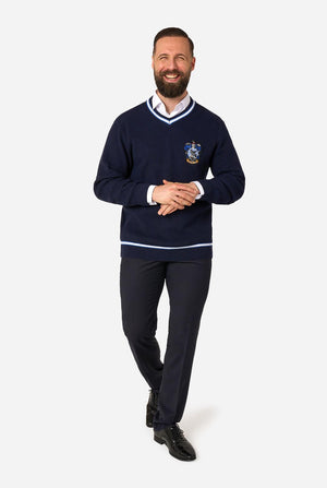 A smiling man wearing a navy blue v-neck Harry Potter Ravenclaw sweater with white trim and a school-style crest on the chest, paired with dark trousers and black dress shoes, standing against a light gray background.
