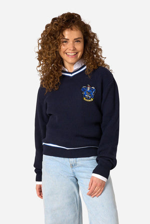 A woman with curly brown hair wearing a navy blue knitted sweater with a Harry Potter Ravenclaw crest on the chest, styled with light blue jeans against a plain white background.