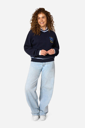 A woman with curly brown hair stands against a light gray background wearing a navy blue Harry Potter Ravenclaw knitted sweater with white stripes and a crest, light-wash wide-leg jeans, and white sneakers.