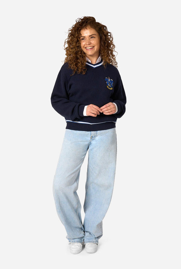 A woman with curly brown hair stands against a light gray background wearing a navy blue Harry Potter Ravenclaw knitted sweater with white stripes and a crest, light-wash wide-leg jeans, and white sneakers.