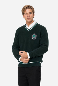 A man wearing a dark green knit v-neck Harry Potter Slytherin sweater with white and light green trim, featuring a Slytherin crest embroidered on the left chest, layered over a white collared shirt and paired with black trousers, set against a plain light gray background.