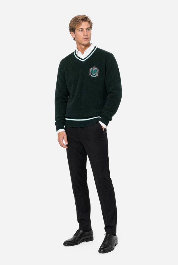 A man modeling a dark green knit Harry Potter Slytherin sweater with white trim and a crest on the chest, paired with black trousers and black shoes, against a light gray background.