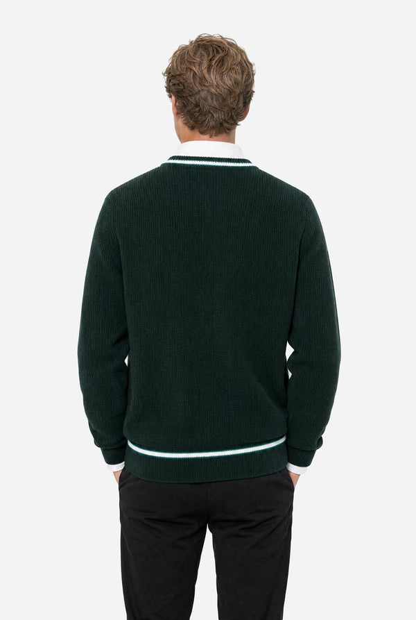 A man viewed from behind wearing a dark forest green ribbed knit sweater with white contrast trim at the collar and hem, paired with black trousers, against a light gray studio background.