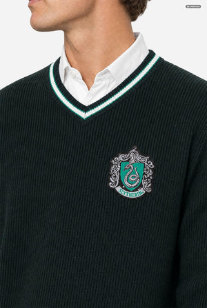 A man wearing a dark green knit v-neck Harry Potter sweater with white and light green trim, featuring a Slytherin crest on the chest, layered over a white collared shirt, set against a plain light gray background. Chest close-up.