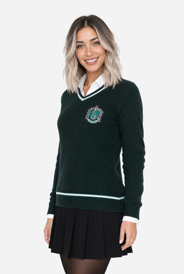 A woman modeling a dark green v-neck knit Harry Potter Slytherin sweater with white and light green trim, featuring a Slytherin crest embroidered on the chest, styled over a white collared shirt and black pleated skirt.