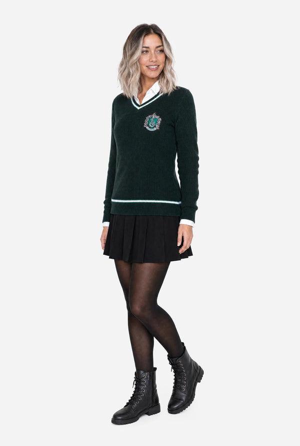 A woman modeling a dark green long-sleeve V-neck Harry Potter Slytherin sweater with white stripes and a crest emblem on the chest, paired with a black pleated skirt, black tights, and black boots, against a light gray background.