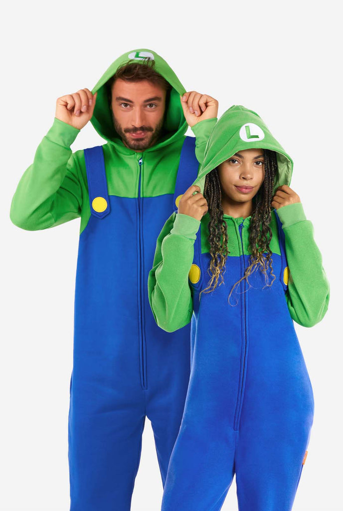 A man and a woman are dressed in a Luigi-themed onesie. The outfit looks as a bright green hoodie with an "L" symbol on the hood and blue overalls with large yellow buttons on the straps. The overall design mimics the look of the popular video game character Luigi, with contrasting green and blue colors. The background is plain, putting focus on the costume.