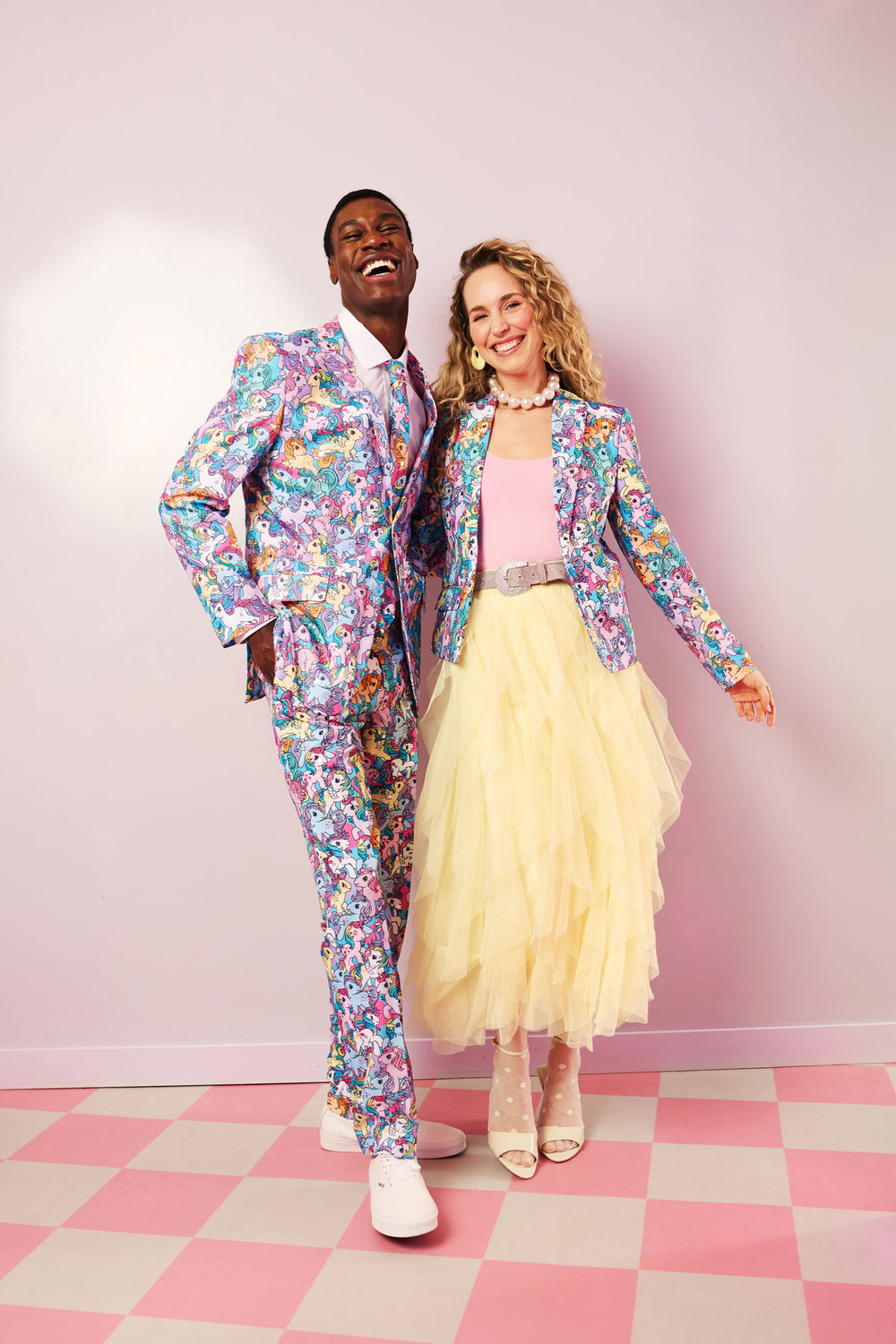 A man in a colorful My Little Pony suit and a woman in a My Little Pony blazer and yellow skirt standing on a checkered floor with a pink wall.