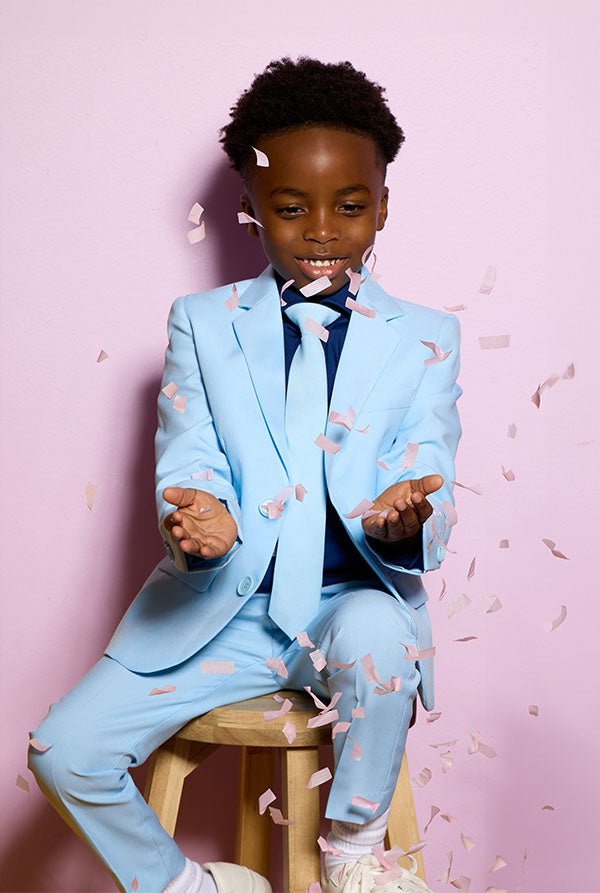 Child in a light blue suit sitting on a stool against a pink background.