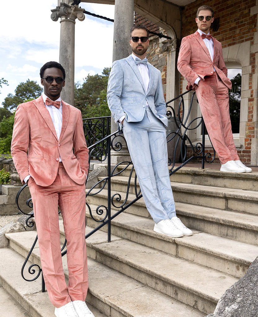 3 men standing outside on stairs wearing velvet tuxedos. The left and right men wearing velvet pink tuxedo and the middle man wearing velvet blue tuxedo. 