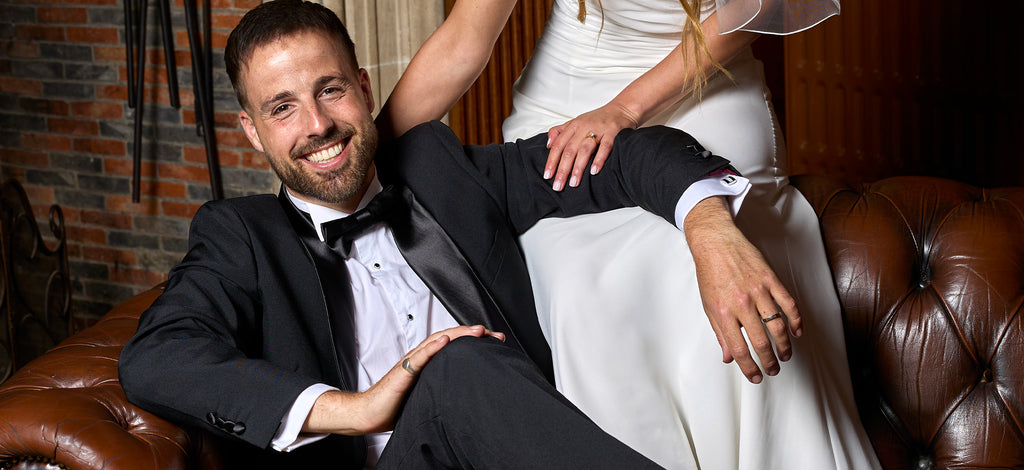 Man sitting on sofa wearing black tuxedo and white shirt. Woman sitting on backrest wearing white dress. Wedding setting.