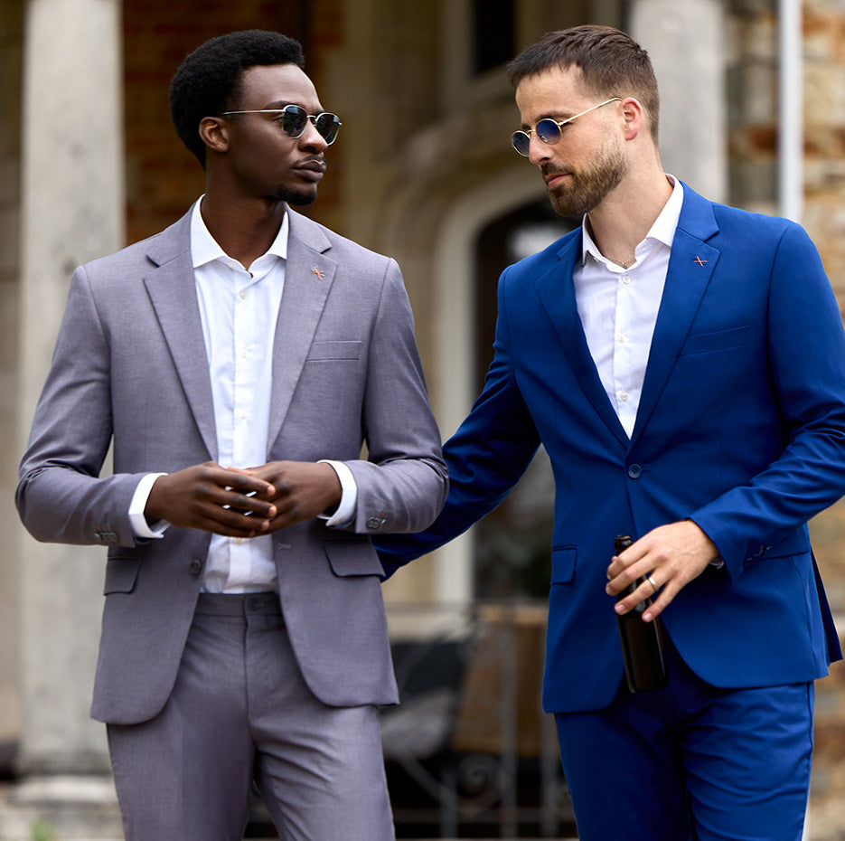 2 men standing outside wearing sunglasses and mens suits. Left man is wearing grey suit with white shirt and right man is wearing dark blue mens suit with white shirt.