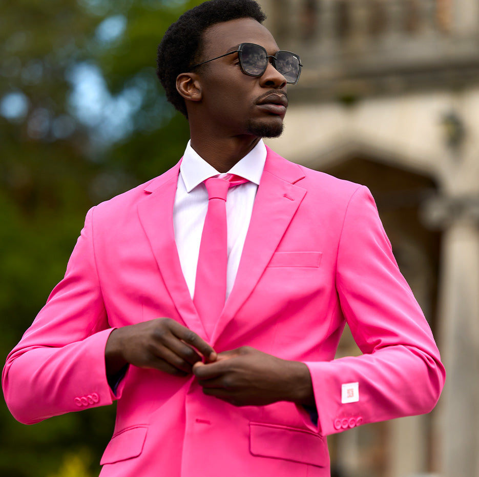 Man standing outside wearing pink mens suit, white shirt and sunglasses. Closing one button of the jacket.