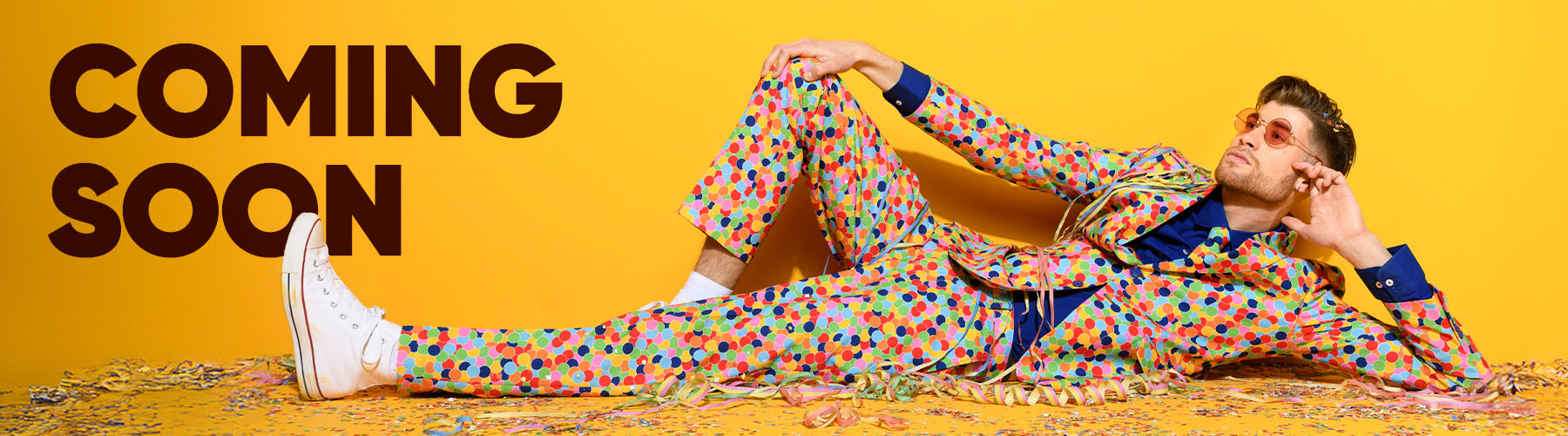 Image with yellow background and a man laying on the floor wearing a confetti suit, and the text Coming Soon on the left side .