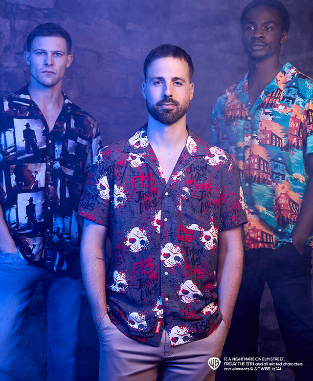 The image features three men standing in front of a stone wall in a dimly lit, misty environment. Each of the men is wearing a different patterned shirt, reflecting a dark and moody vibe. The man on the left wears a shirt with a black and white design of city buildings, the man in the center has a red shirt with skull prints, and the man on the right wears a vibrant turquoise shirt with a tropical pattern. The lighting casts a blue hue over the scene, creating an edgy and atmospheric feel, suggesting an eve