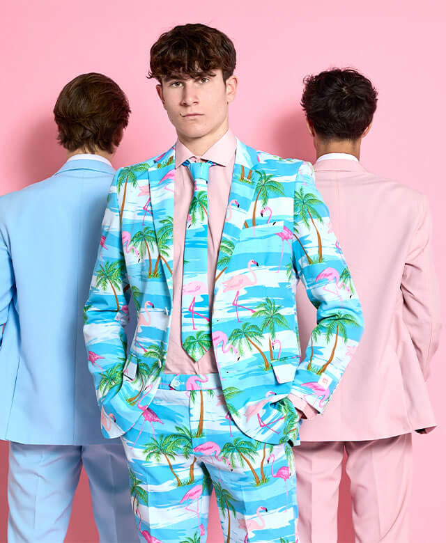 The image shows three men wearing suits in a bright, pink-colored room. The man in the foreground is facing forward and wearing a tropical-themed suit with palm trees, flamingos, and water scenes in pastel colors of blue, green, and pink. He has his hands in his pockets and is looking directly at the camera. Behind him are two men, one wearing a light blue suit and the other in a pastel pink suit. Both have their backs turned to the camera, further emphasizing the unique, playful, and summer-like style of t