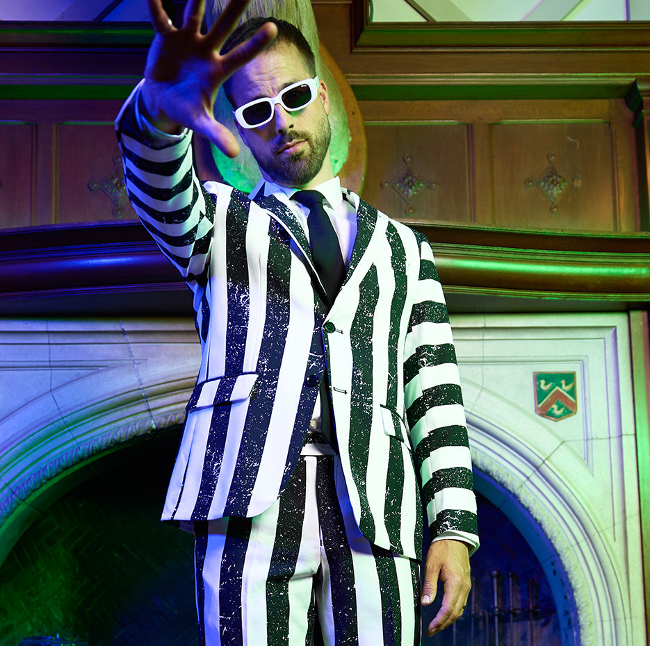 A vibrant promotional image for Beetlejuice. The background is a mix of bright green and purple colors. In the center, a man dressed in a bold black-and-white striped suit, resembling Beetlejuice. He is wearing sunglasses. 