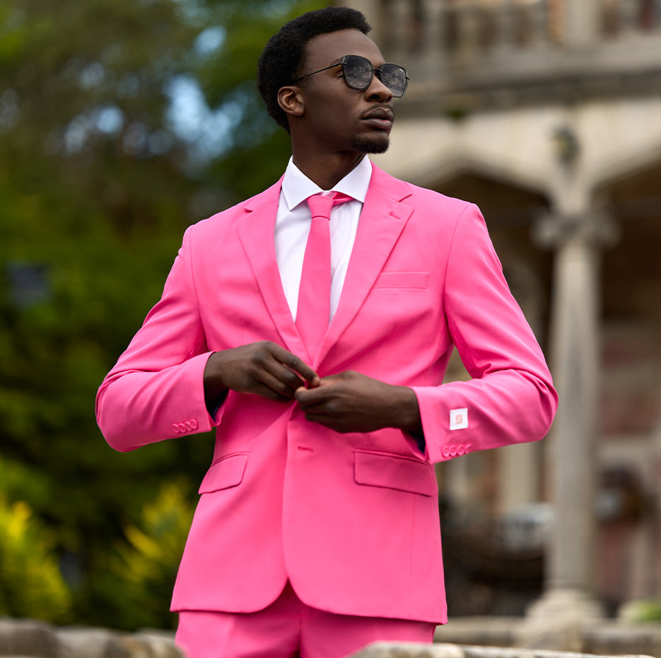 Man standing outdoors wearing a pink mens suit and a white shirt. Tying one button of the jacket.