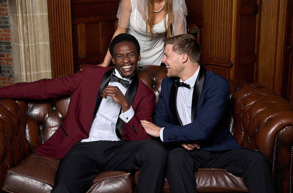 2 men sitting on a brown sofa laughing. The left man is wearing a tuxedo with a burgundy jacket and black pants and bow tie, the right man is wearing a tuxedo with a dark blue jacket and black pants and bow tie. Behind the sofa a woman wearing a with dress.