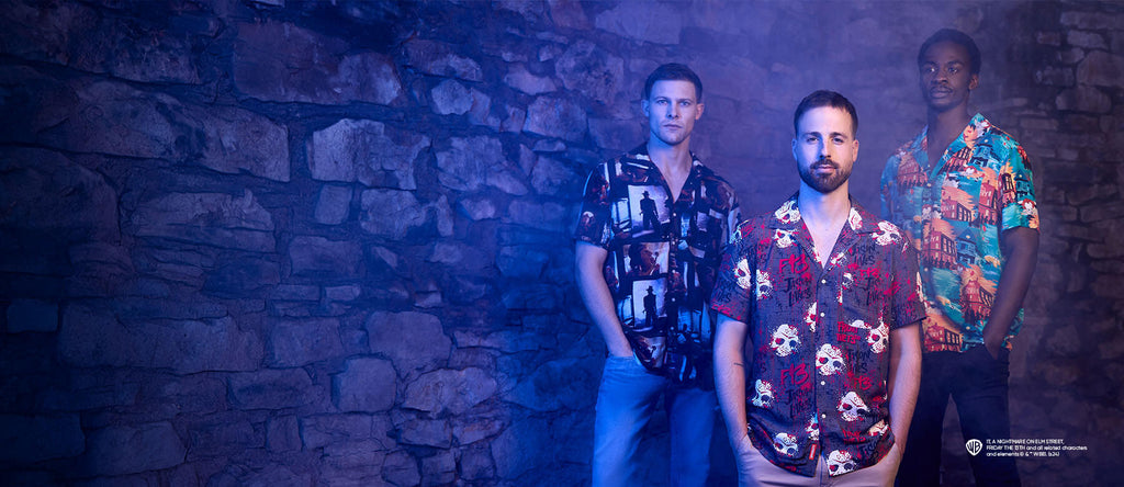 The image features three men standing in front of a stone wall in a dimly lit, misty environment. Each of the men is wearing a different patterned shirt, reflecting a dark and moody vibe. The man on the left wears a shirt with a black and white design of city buildings, the man in the center has a red shirt with skull prints, and the man on the right wears a vibrant turquoise shirt with a tropical pattern. The lighting casts a blue hue over the scene, creating an edgy and atmospheric feel, suggesting an eve