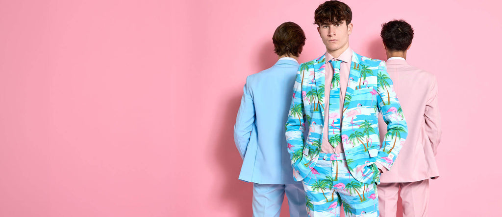 The image shows three men wearing suits in a bright, pink-colored room. The man in the foreground is facing forward and wearing a tropical-themed suit with palm trees, flamingos, and water scenes in pastel colors of blue, green, and pink. He has his hands in his pockets and is looking directly at the camera. Behind him are two men, one wearing a light blue suit and the other in a pastel pink suit. Both have their backs turned to the camera, further emphasizing the unique, playful, and summer-like style of t