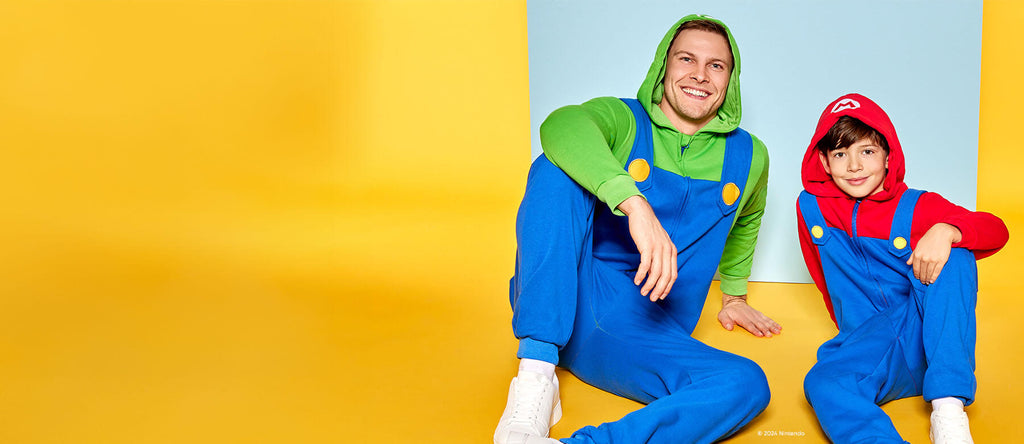 The image shows two people seated on the floor against a vibrant yellow background. One is an adult, and the other is a child. The adult is dressed as Luigi, from the Mario video game series, wearing a green hoodie, blue overalls, and white shoes. The child is dressed as Mario, wearing a red hoodie, blue overalls, and white shoes. Both are smiling and sitting with their legs bent in a relaxed position. Behind them, there is a light blue square that contrasts with the yellow background, further drawing atten