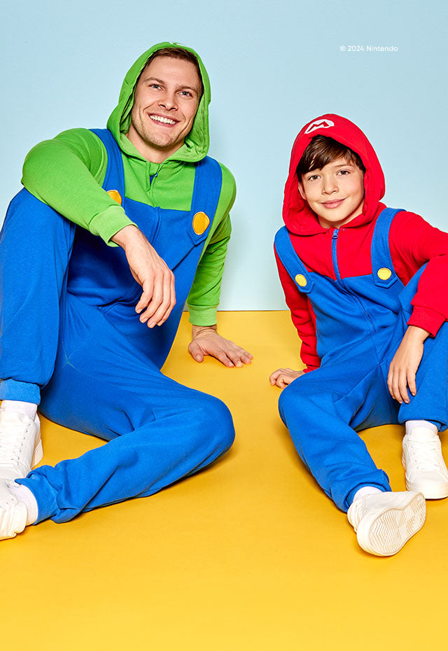 Man and kid sitting on the ground with one foot flat on the floor and the elbow on the knee.  Background is yellow and blue screen behind them. Man is wearing green and blue luigi themed onesie and the kid is wearing a red and blue Super Mario themed onesie.