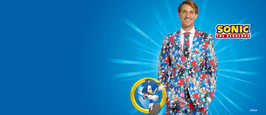 The image shows a man standing in front of a blue background. He is wearing a suit that features a vibrant and detailed pattern based on the character Sonic the Hedgehog. The suit's design includes images of Sonic, as well as other characters and symbols from the Sonic the Hedgehog franchise. The man is smiling and facing the camera, and he is wearing a white shirt with a red tie underneath the patterned suit.
On the right-hand side of the image, there is a circular emblem featuring Sonic the Hedgehog runni