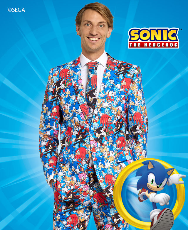 The image shows a man standing in front of a blue background. He is wearing a suit that features a vibrant and detailed pattern based on the character Sonic the Hedgehog. The suit's design includes images of Sonic, as well as other characters and symbols from the Sonic the Hedgehog franchise. The man is smiling and facing the camera, and he is wearing a white shirt with a red tie underneath the patterned suit.
On the left side of the image, there is a circular emblem featuring Sonic the Hedgehog running.