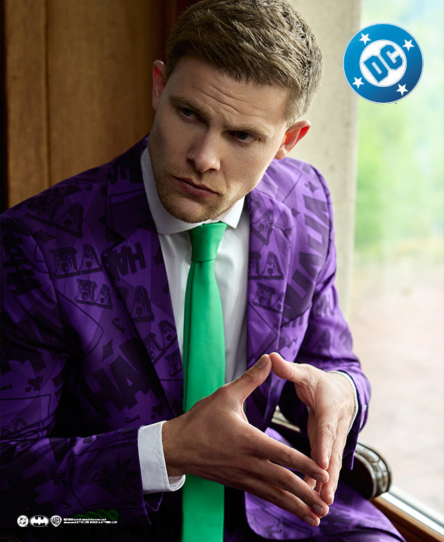 A man sitting on a chair in front of a window, wearing a purple mens suit with a The Joker theme. Also wearing a white shirt and a green  tie. Close up on the upper part of his body.