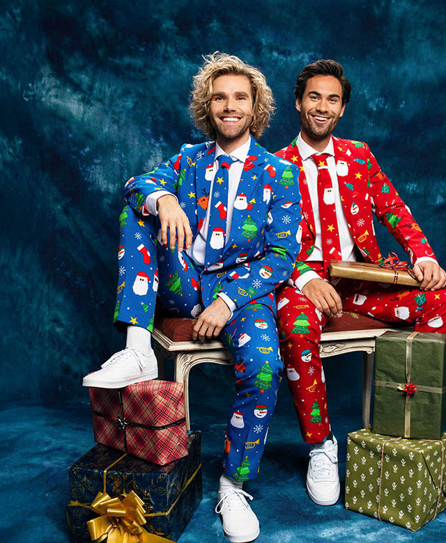 Two man sitting on a bench in fornt of a blue curtain wearing a Christmas suit. Left man wearing a blue Christmas suit and the right man wearing red Christmas suit.