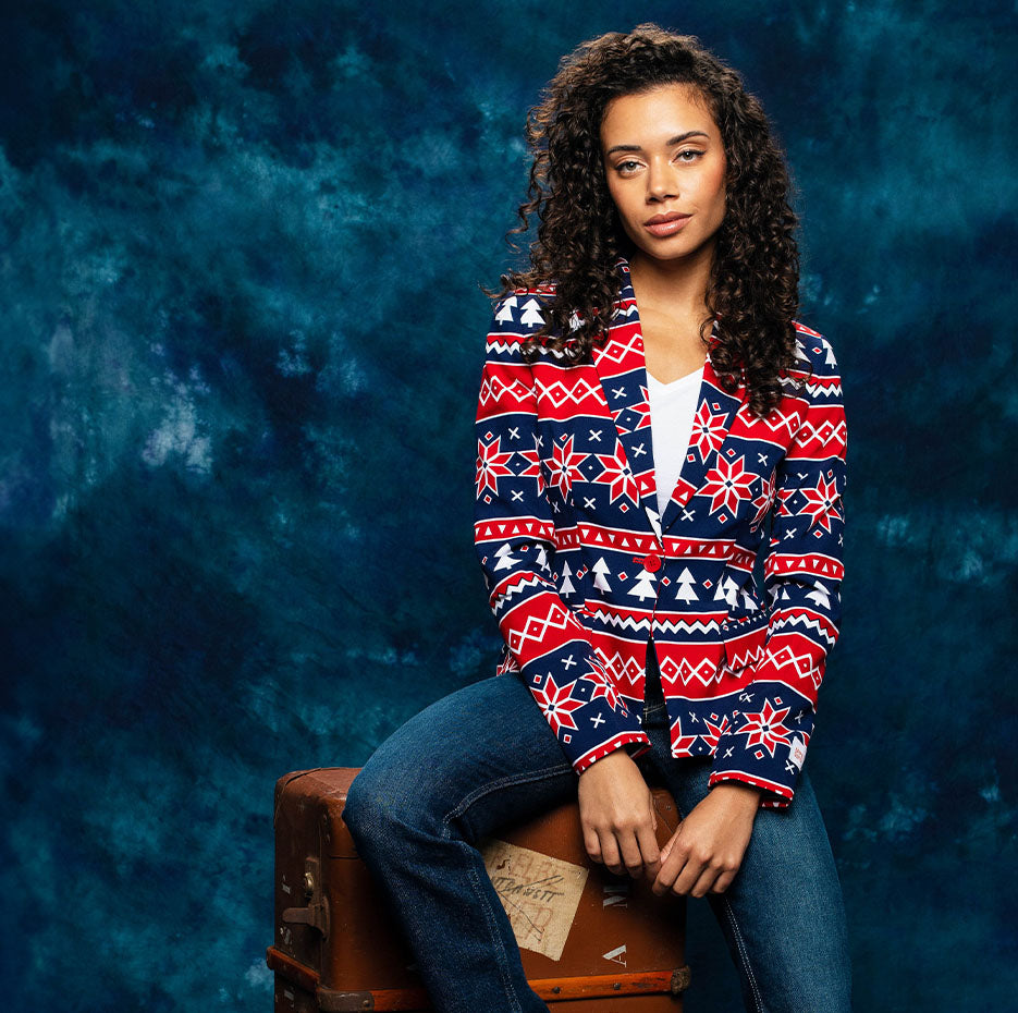 Image with blue background and a woman sitting on a suitcase wearing a bleu and red Nordic Christmas blazer.