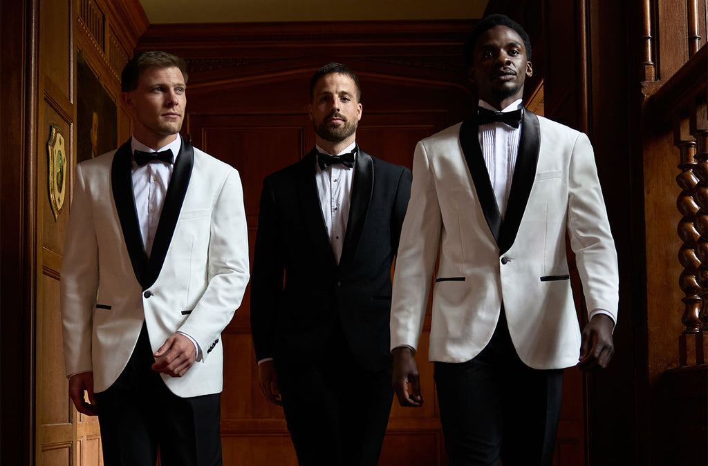 3 man in a hallway wearing tuxedos. 1 man wearing a black tuxedo and 2 men wearing a tuxedo with a white jacket and black pants.