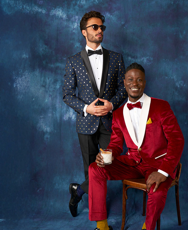 Image with blue background and 2 men wearing Christmas tuxedos. 1 man wearing red velvet tuxedo and a man wearing blue tuxedo with golden dots.