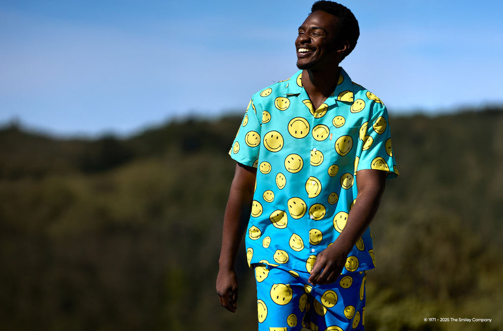 Image of man walking outside in a forest like environment wearing a blue summer set with Smiley print. 