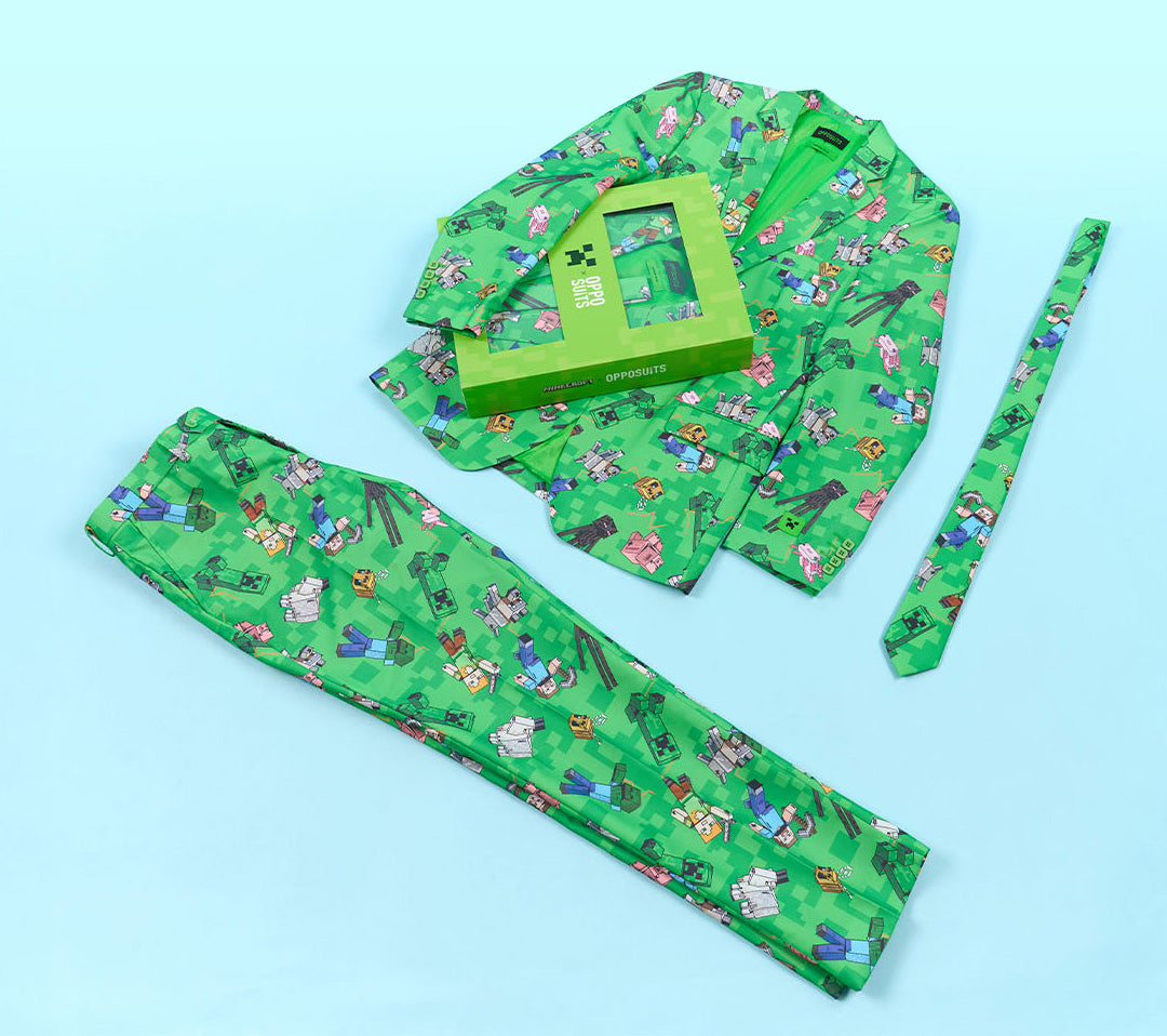 A green Minecraft-themed matching suit laid out on a light blue background, including a long-sleeve blazer, pants, a tie, and a small gift box. The fabric has an all-over Minecraft-style character print in multiple colors.