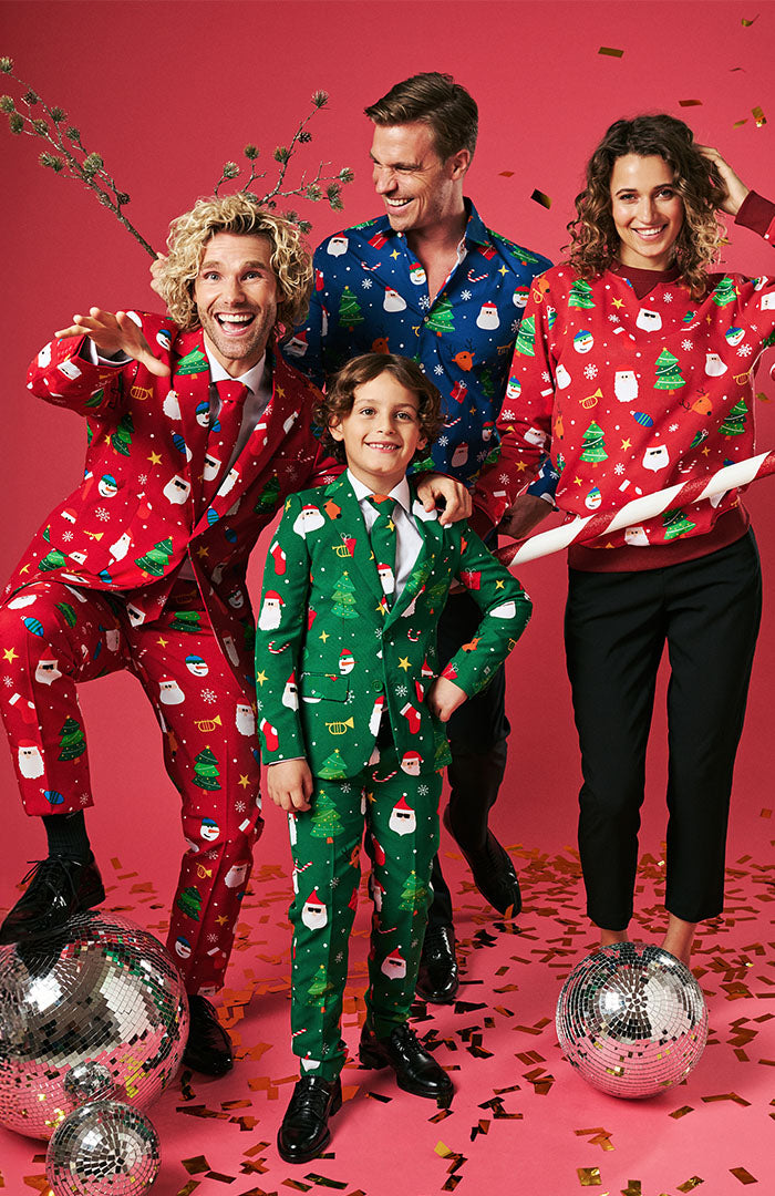 Image with red background and 4 persons on it, 2 men, 1 woman and one kid. The man on the left is wearing a red Christmas suit. The man in the back is wearing a blue Christmas shirt. The woman on the right is wearing a red Christmas Sweater and the kid in fornt is wearing a green Christmas suit.