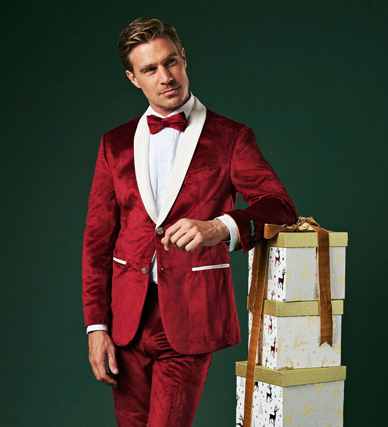 Image with green background and man wearing a red and white velvet Christmas Tuxedo and he is leaning on a stack of presents.
