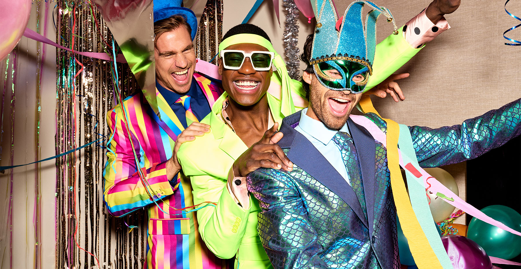 Image with 3 men partying and doing the polonaise. Balloons and garlands on the background. The man in front wearing a green/blue shiny suit, the middle man is wearing a neon green suit, and the man in the back a rainbow colored suit.