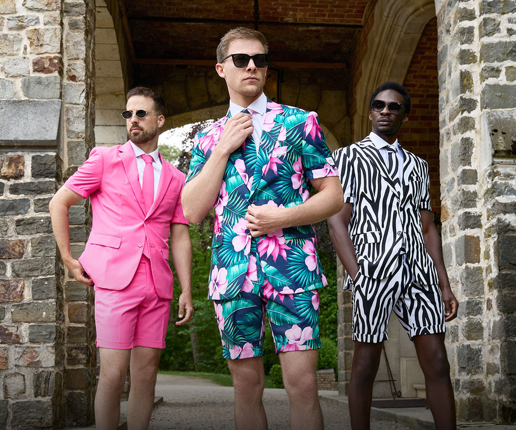 Image of 3 men standing in front a stone wall wearing summer suits consisting of short, short sleeve blazer and tie. One wearing a pink suit, one a zebra striped suit and one a flower print suit.