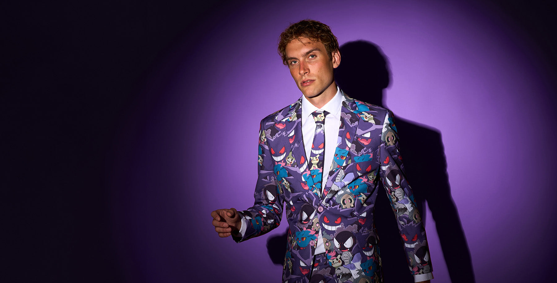 A man wearing a purple suit jacket with a colorful cartoon character print, a white dress shirt, and a patterned tie, standing against a purple spotlight background with a dark shadow.