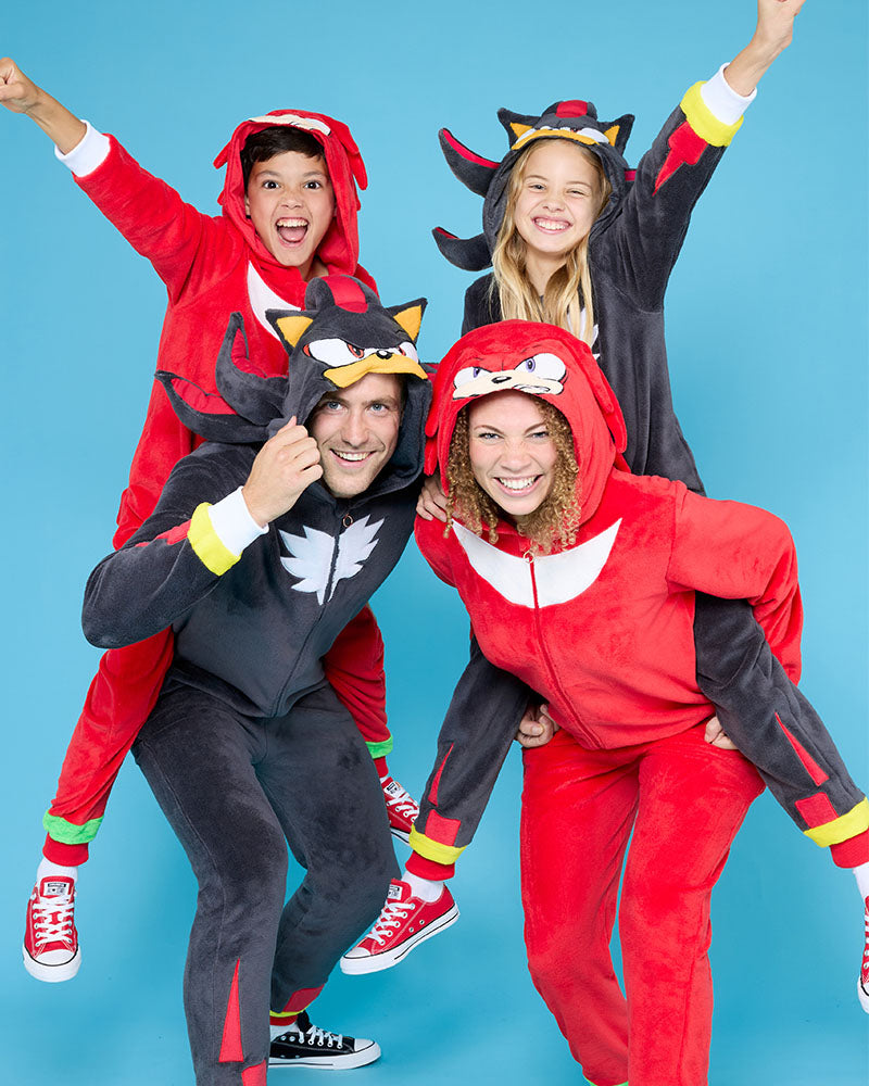 A family of four posing in matching superhero-style hooded onesies against a bright blue studio background. The set includes a  black Shadow themed one-piece worn by the father, a red Knuckles one-piece worn by the mother, and matching dark gray/black and red onesies worn by the two children.