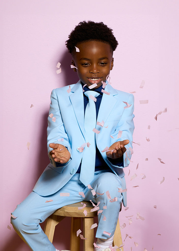 Kid sitting on stool in front of pink background. Kid is wearing a light blue boys suit and a dark blue shirt. He is holing his hands in front a him catching confetti.