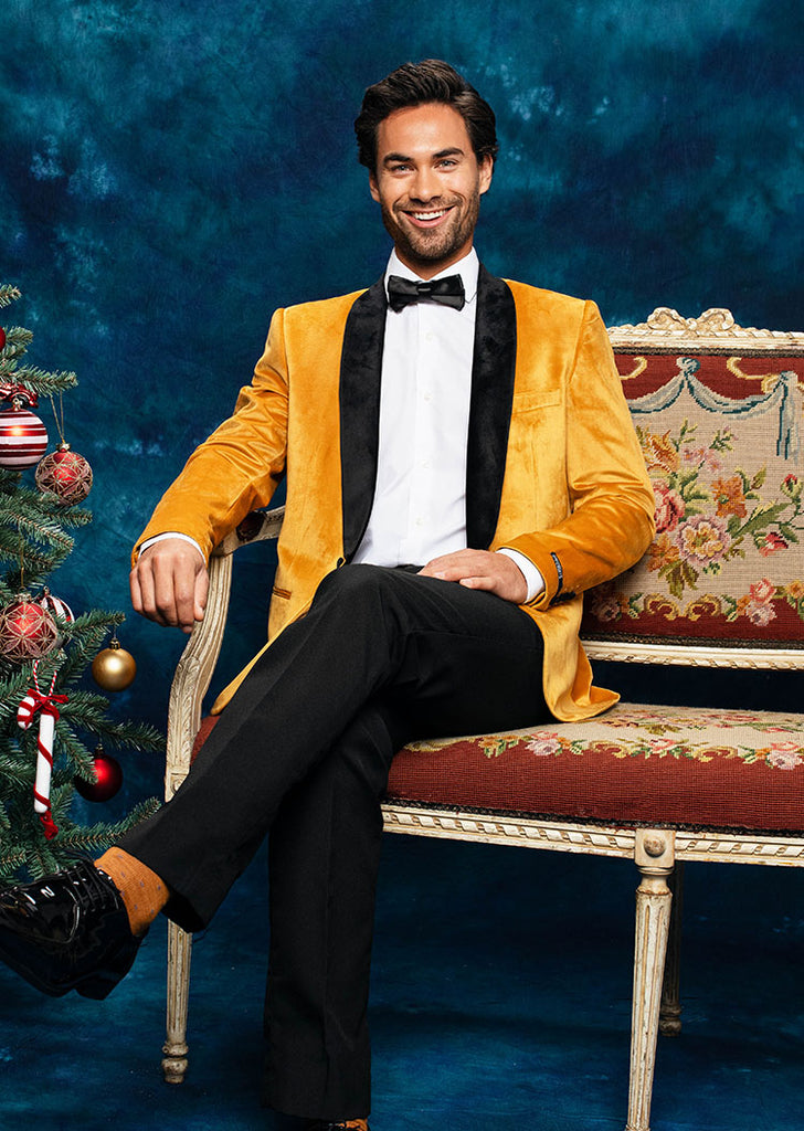 Image with a blue background and a man on a sofa wearing a golden velvet blazer with a black satin lapel, and a black pants.